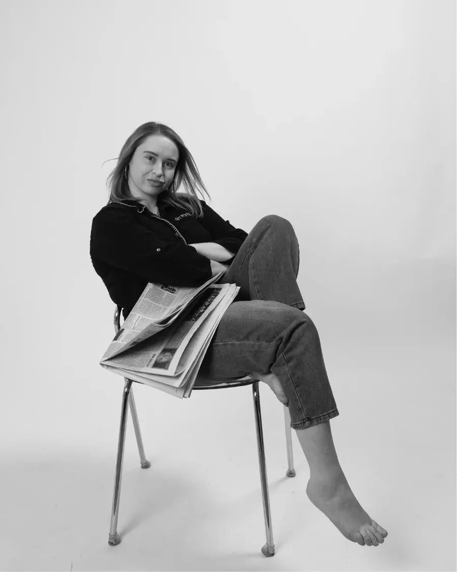 Emily sitting sideways on a chrome chair, arms crossed, newspapers on her lap