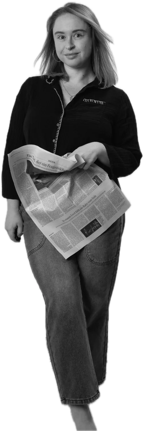 Emily standing barefoot, holding a crumpled German newspaper and smiling slightly
