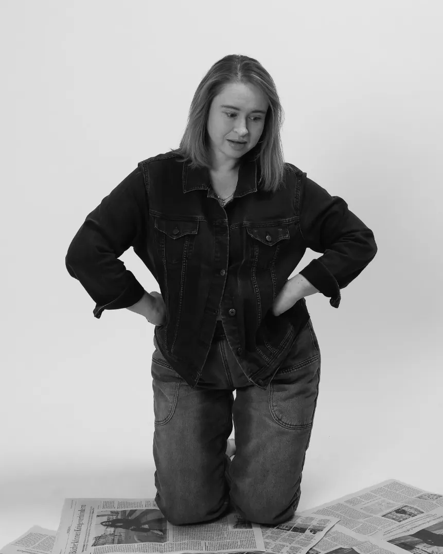 Emily kneeling on newspapers in a denim jacket, hands on hips, frowning down at the headlines