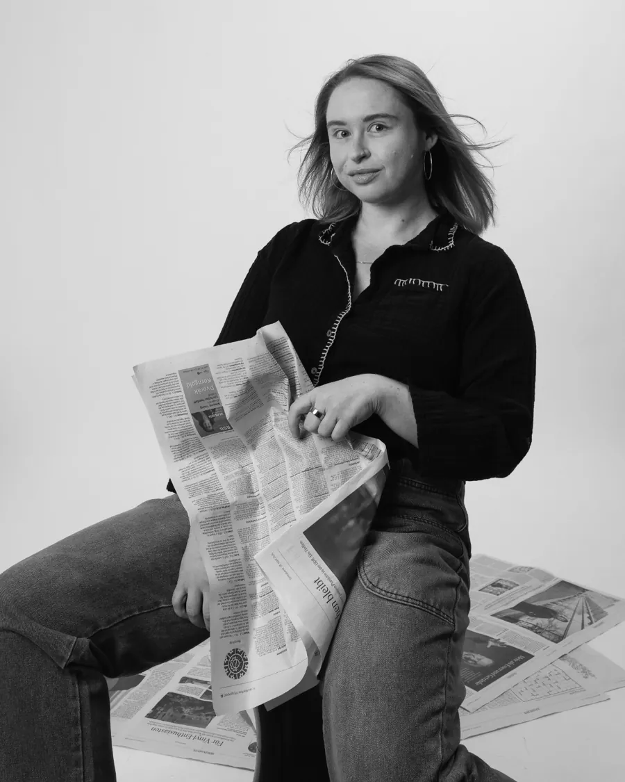 Emily sitting with a newspaper across her lap, giving the camera a knowing look